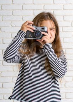 little girl in a striped blouse with a retro camera photographing standing near a light brick wall