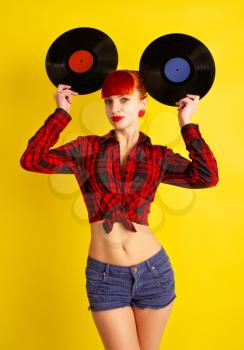 Red-haired girl dressed in retro style in a red checkered shirt and shorts does not have attached vinyl records to her head like ears on a yellow background.