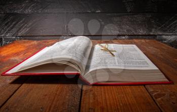 An open holy bible with a red cover and a small metal cross on a chain on an old dark wooden surface