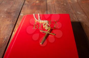 closed holy bible with a red cover and a small metal cross on a chain on an old dark wooden surface