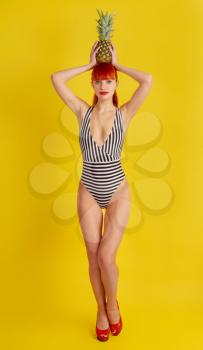 A young girl in a retro striped bathing suit and high-heeled shoes put a pineapple on her head on a bright yellow background