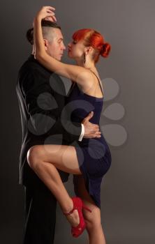 A man in a strict dark suit and a beautiful girl in a blue dress and heeled shoes dance a slow sensual dance on a dark background.