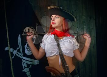 Young attractive armed girl pirate captain looks into the distance on the background of the flag Jolly Roger