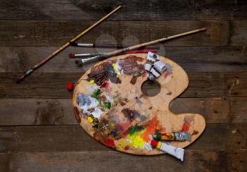 wooden classic palette smeared with multi-colored paints lying on a wooden background next to used brushes and tubes of paint