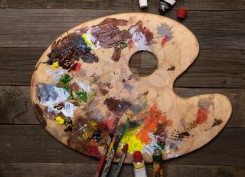 wooden classic palette smeared with multi-colored paints lying on a wooden background next to used brushes and tubes of paint