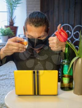 An adult male in a medical mask while in quarantine drinks alcohol while communicating with a friend via the Internet