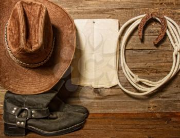 Very rusty old horseshoe symbol of luck and lasso hanging on a wooden wall cowboy hat and boots and blank sheet of paper with place for text.