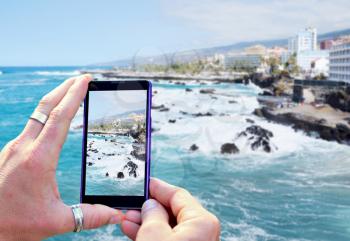 View over the mobile phone display during taking a picture of Puerto de la Cruz coast in Tenerife. Holding the mobile phone in hands and taking a photo. Focused on mobile phone screen. 