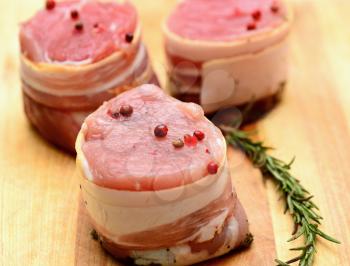 Closeup of fresh raw bacon wrapped pork tenderloin medallions sprinkled with whole red pepper and rosemary.