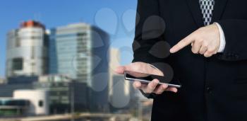 Businessman Holding Smartphone in Hand And Pointing Index Finger At The Phones Screen With Business City and Corporate Buildings In Background