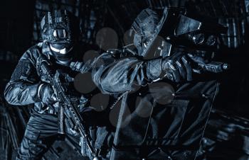 Police special operations tactical group fighters in black uniform and mask, armed pistol and service rifle, protecting themselves with armored ballistic shield, sneaking in darkness during night raid