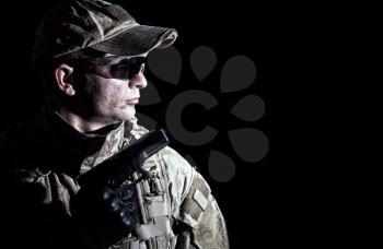Army soldier armed service pistol in camo combat uniform, cap and ballistic goggles, standing in darkness, holding handgun in hand, low key, side view, portrait isolated on black background, copyspace