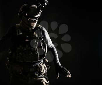 Special operations fighter in helmet with night-vision, thermal imaging device, load carrier carefully moving with caution in darkness, holding hand on pistol, ready for fight during dangerous mission