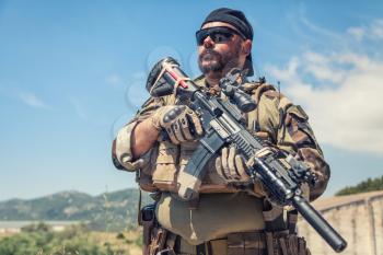 Private military company mercenary, brutal looking special forces fighter in battle uniform and plate carrier, wearing radio headset and sunglasses, holding service rifle in hands, ready to fight