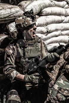 Navy SEALs rifleman, army soldier in combat uniform and helmet, grabbing assault rifle, screaming, yelling while covering behind sandbags. Special forces infantryman attacking enemies in trench