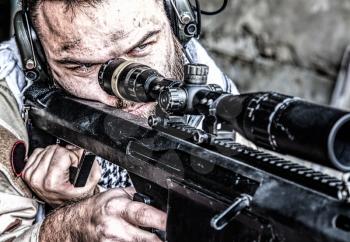 Portrait of U.S. Navy SEAL sniper on firing position, armed with large caliber sniper rifle with telescopic sight, wearing tactical headset with microphone, observing territory, searching targets