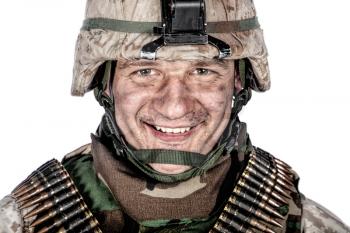Close up studio portrait of friendly looking marine, military veteran with dirt or soot on face in ragged combat helmet and ammo belts over shoulders, looking at camera with smile, cropped on white