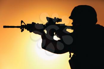 Silhouette of modern army infantry soldier standing on background of setting sun and aiming assault rifle with collimator sight. Special operations rifleman, elite forces shooter shooting in enemy