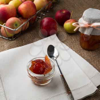 Glass jar of apple jam, spoon, jar of marmalade, linen napkin and apples in metal wire basket on a burlap covered table, square. Selective focus. The toning
