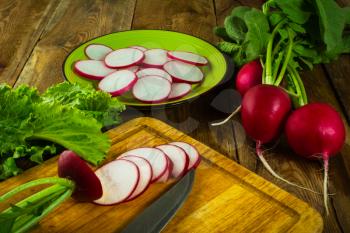 A bunch of fresh radishes, lettuce leaves, slices of radish and knife on old chopping board
