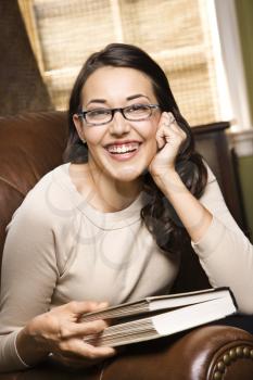 Caucasian/Hispanic young woman sitting in leather chair holding book and smiling at viewer.