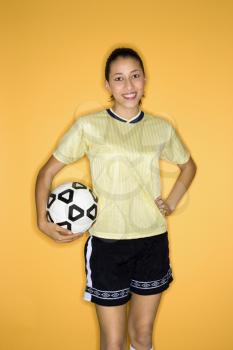 Royalty Free Photo of a Teen Girl Holding a Soccer Ball at Hip Standing Against a Yellow Background