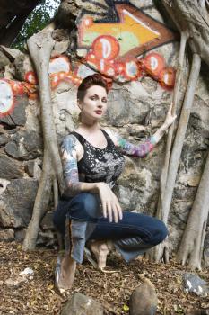 Sexy Caucasian tattooed woman crouching next to wall covered in graffiti and Banyan tree branches.