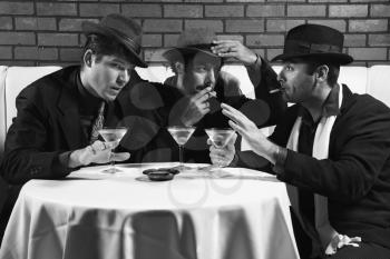 Royalty Free Photo of Three Men Wearing Retro Suits Sitting at a Table Drinking, Smoking and Talking