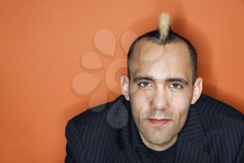 Head and shoulder portrait of Caucasian man with mohawk wearing suit against orange background.