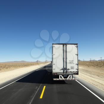Royalty Free Photo of a Back View of a Semi Truck Traveling Down the Highway Towards the Horizon Under a Clear Blue Sky
