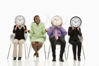 Business people sitting holding clocks over faces while African-American businesswoman shrugs.