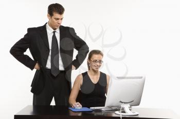 Royalty Free Photo of a Man With Hands on Hips Looking Over Shoulder of a Woman Sitting at Computer