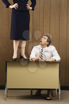 Royalty Free Photo of a Male Sitting at a Desk Looking Up To a Woman Standing on a Desk