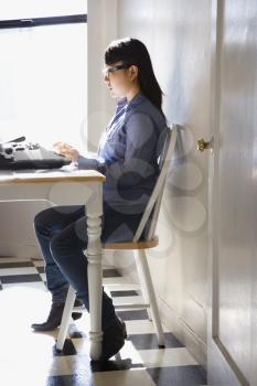 Pretty young Asian woman sitting in chair typing on typewriter in kitchen.