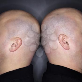 Close up of Caucasian mid adult identical twin bald men standing back to back.