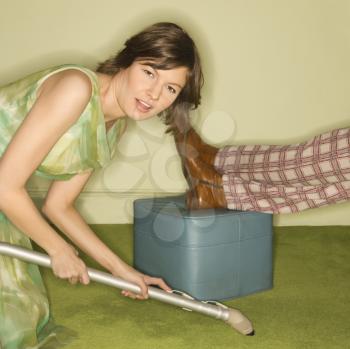 Royalty Free Photo of an Unhappy Woman Kneeling and Vacuuming Carpet Around a Male Feet Resting on Foot Stool