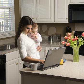 Caucasian woman holding baby  and typing on laptop computer in kitchen.