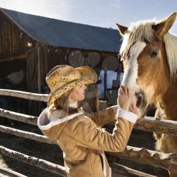 Young Caucasian woman petting horse with stable in background.