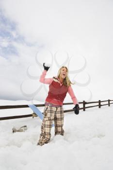Young woman in winter clothes by fence in snowy field laughing and ready to throw snowball.