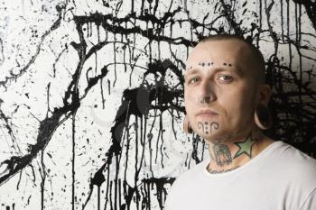 Royalty Free Photo of a Tattooed and Pierced Man Leaning Against a Paint Splattered Background