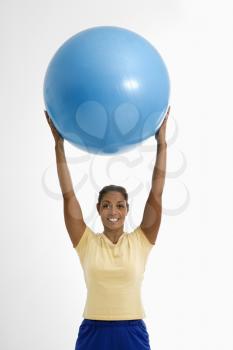 Mid adult multiethnic woman standing and holding blue exercise ball over her head looking at viewer and smiling.