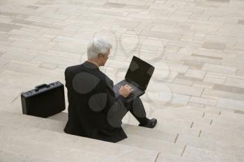 Royalty Free Photo of a Back View of a Businessman Sitting on Steps Outdoors With a Laptop and Briefcase