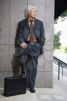 Caucasian middle aged businessman leaning on wall outdoors with foot on briefcase.