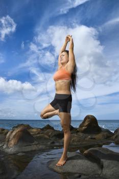 Asian woman on rock by ocean in Maui, Hawaii doing standing tree yoga pose
