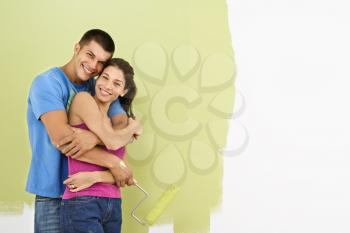 Royalty Free Photo of a Couple Smiling and Embracing in Front of a Partially Painted Wall Holding a Paintbrush and Roller