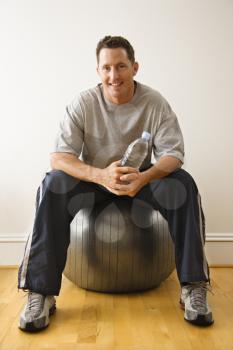 Royalty Free Photo of a Man Holding a Water Bottle Sitting on a Balance Ball at a Gym Smiling