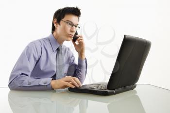 Asian Royalty Free Photo of a Businessman Sitting at a Desk Working on a Laptop Talking on a Cellphone