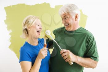 Middle-aged couple painting wall green playing with paint utensils.