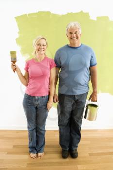 Portrait of happy adult couple standing in front of half-painted wall with paint supplies 'American Gothic' style.