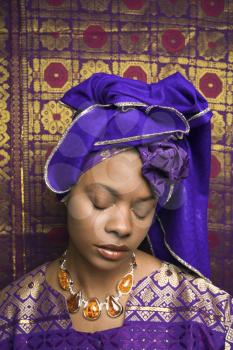 Portrait of an African American woman wearing traditional African clothing and closing her eyes in front of a patterned wall. Vertical format.